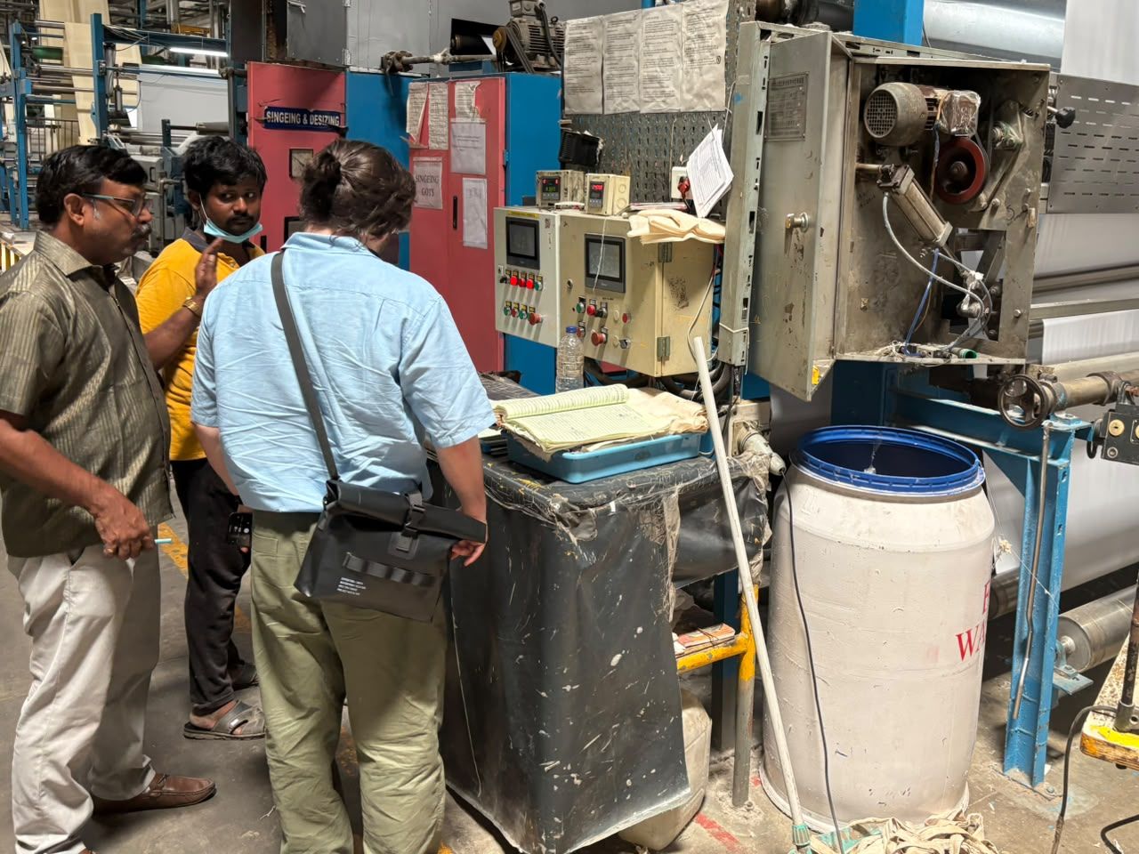 Rhys with two machine operators at a finishing line control panel, a handwritten production log open on the bench